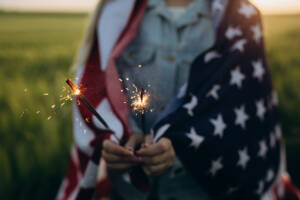 A person wrapped in an American flag holding two sparklers outdoors.