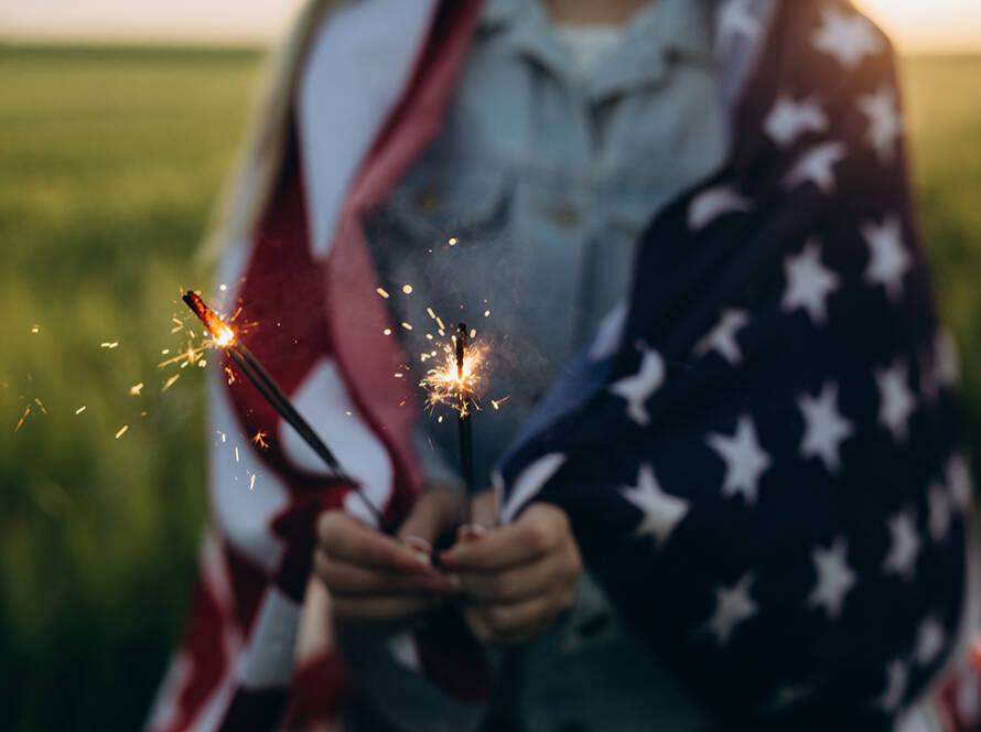 A person wrapped in an American flag holding two sparklers outdoors.